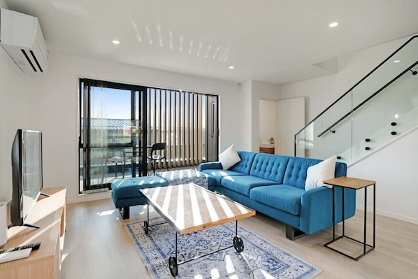 A modern living room with a sofa, ottoman, and coffee table, a television, and a staircase.