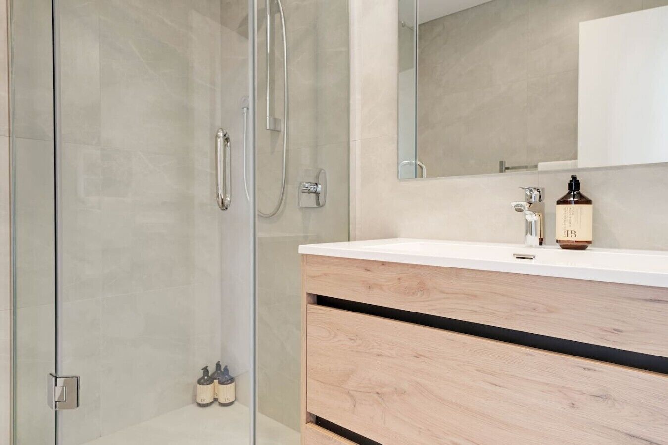 A modern bathroom with a glass shower enclosure, a white vanity with a wooden drawer, and a bottle of lotion on the counter.