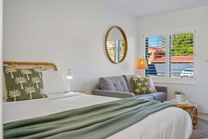 A bedroom with a bed, a grey sofa, a round side table, a mirror, and a window with blinds.