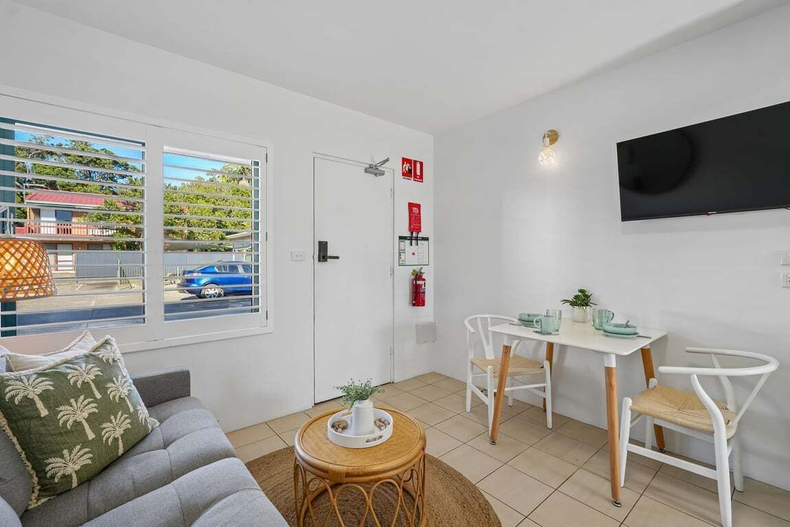 A modern living room with a sofa, a dining table with chairs, a TV mounted on the wall, and a window with blinds.