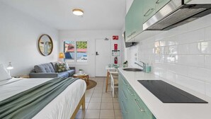 A compact room with a bed, a small seating area, a kitchenette with white cabinets, and a window with a view. - Murrays Beach Studio (Sawtell)