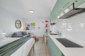 A compact room with a bed, a small seating area, a kitchenette with white cabinets, and a window with a view. - Murrays Beach Studio (Sawtell)