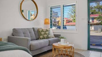 A modern living room with a grey sofa, a round wooden coffee table, a mirror, and a window with blinds.