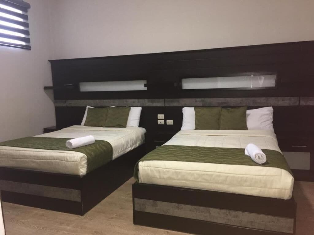 Dos camas individuales con ropa de cama verde, un cabecero oscuro y una ventana con persianas.