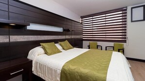 A hotel room with a bed, bedside tables, a window with blinds, and a small seating area.