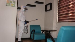 Una persona con traje protector blanco usando un atomizador en una habitación con sillas color turquesa y una mesa.