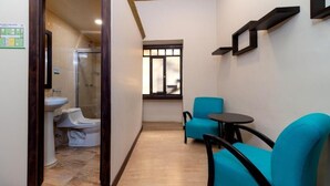 A narrow hallway with a bathroom on the left, a window with blinds, and a seating area with two chairs and a small table.