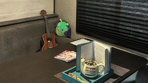 Een kamer met een houten vloer, een gitaar, een groen pluche speelgoed, een raam met zonwering, en een tafel met een decoratief object en een notitieboek.