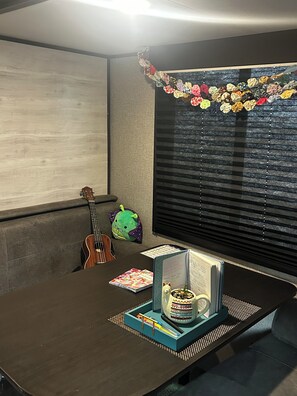 A room with a wooden floor, a guitar, a green plush toy, a window with blinds, and a table with a decorative item and a notebook. - 2-bedroom RV camper in beautiful Huntsville located in Hobbs Island by River (Huntsville)