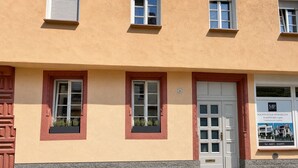 Un bâtiment à la façade beige, doté de deux fenêtres avec volets, d’une porte et d’un petit panneau indiquant les coordonnées.