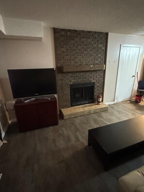 Ein Wohnzimmer mit Kamin aus Backsteinen, einem Flachbildfernseher, einem roten Pouf und einem dunklen Couchtisch.