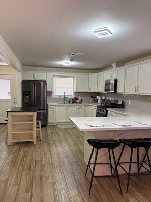 A modern kitchen with white cabinets, stainless steel appliances, a wooden bar stool, and a marble countertop.