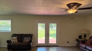 Un salón con un sofá de cuero marrón, un ventilador de techo y vistas a un área exterior verde a través de una puerta de vidrio.