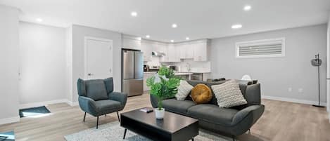 A modern living room with a sofa, armchair, and coffee table, a kitchen in the background, and a dining area.