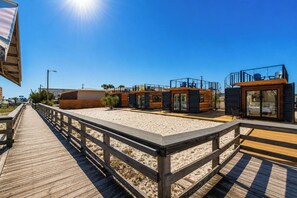 Un muelle de madera que conduce a una propiedad frente a la playa con varias cabañas y una sala con paredes de vidrio.