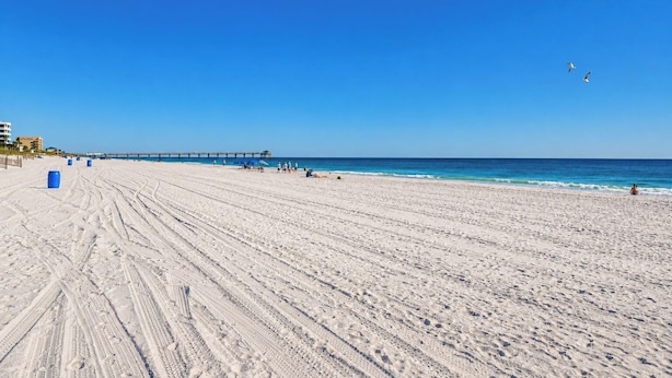 Una playa extensa y arenosa con un muelle que se adentra en el agua, cielos despejados y algunas personas a lo lejos.
