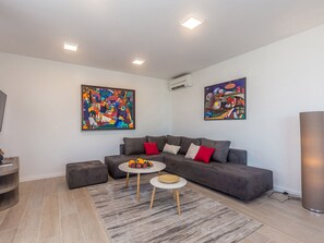 Ein modernes Wohnzimmer mit einem grauen Sofa, zwei Beistelltischen und einer Obstschale. An den Wänden befinden sich zwei abstrakte Gemälde und eine Klimaanlage.