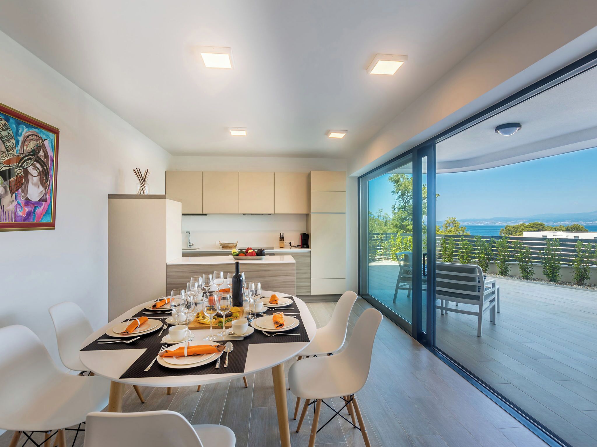 A modern dining area with a table set for four, a kitchen in the background, and a large glass door leading to an outdoor terrace with a view.