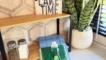 Une étagère sur laquelle on peut lire l’inscription « TEMPS DE DÉTENTE AU LAC », une bouteille verte de liquide vaisselle Palmolive, deux salières et un pot contenant une plante serpent.