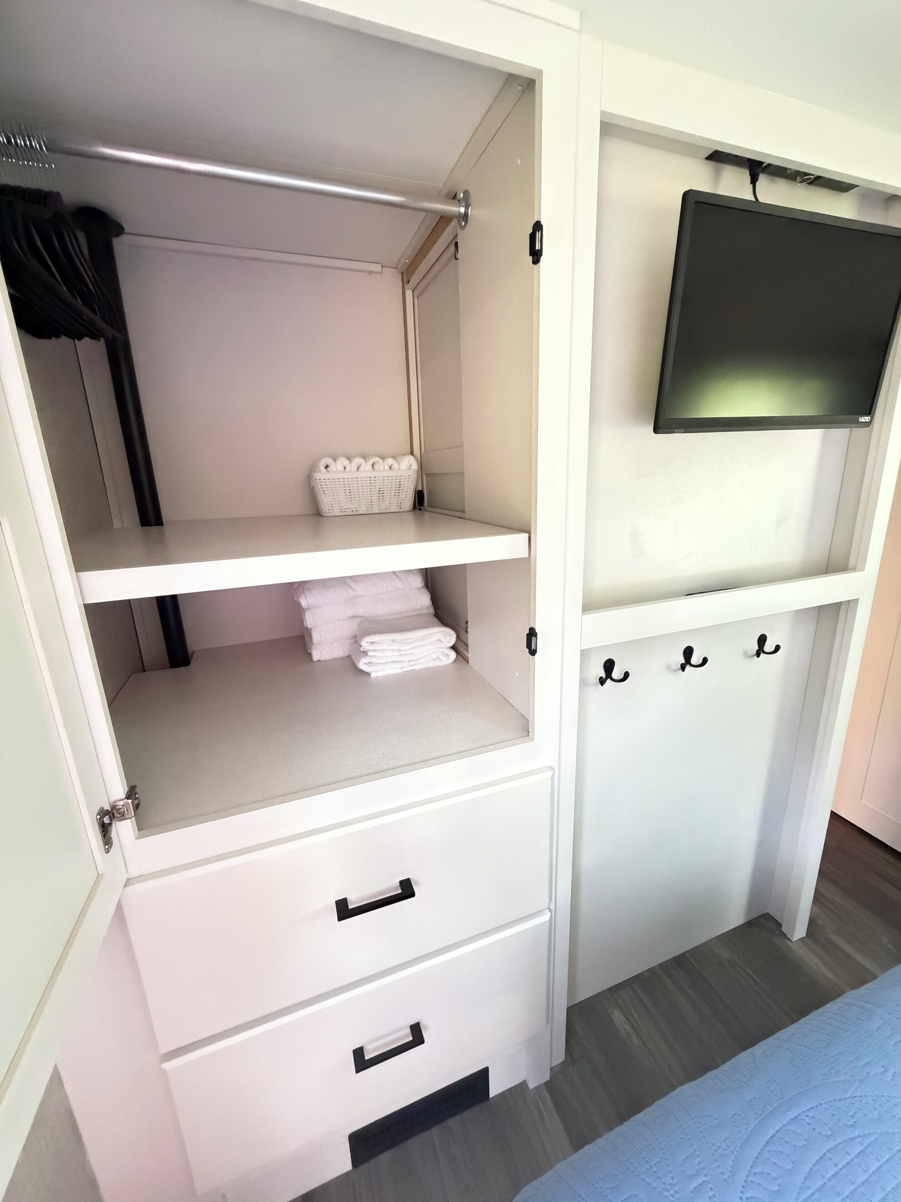 A white wardrobe with a flat-screen TV mounted above it, a hanging rod, and a shelf with folded towels.