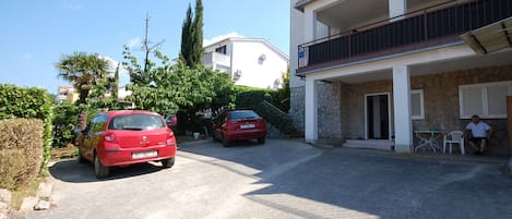 A residential street with two parked cars, a stone building with a balcony, and a clear sky.