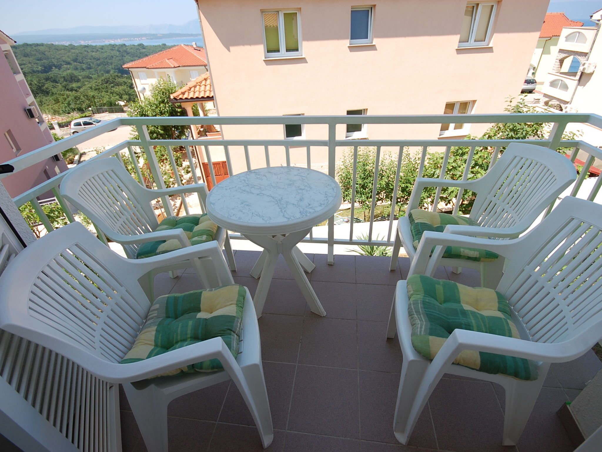 A balcony with a round table and four white plastic chairs, each with a green cushion.