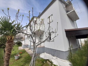 Un edificio de dos pisos con fachada blanca, un balcón en el nivel superior y un jardín con una palmera y una planta en maceta.