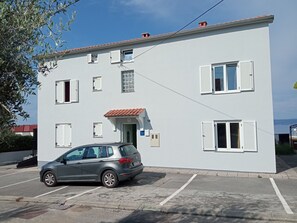A two-story building with white walls and red-tiled roof, a parked car, and a small tree in the foreground. - Apartment Fejer - 4 by Interhome (Krk/Njivice)