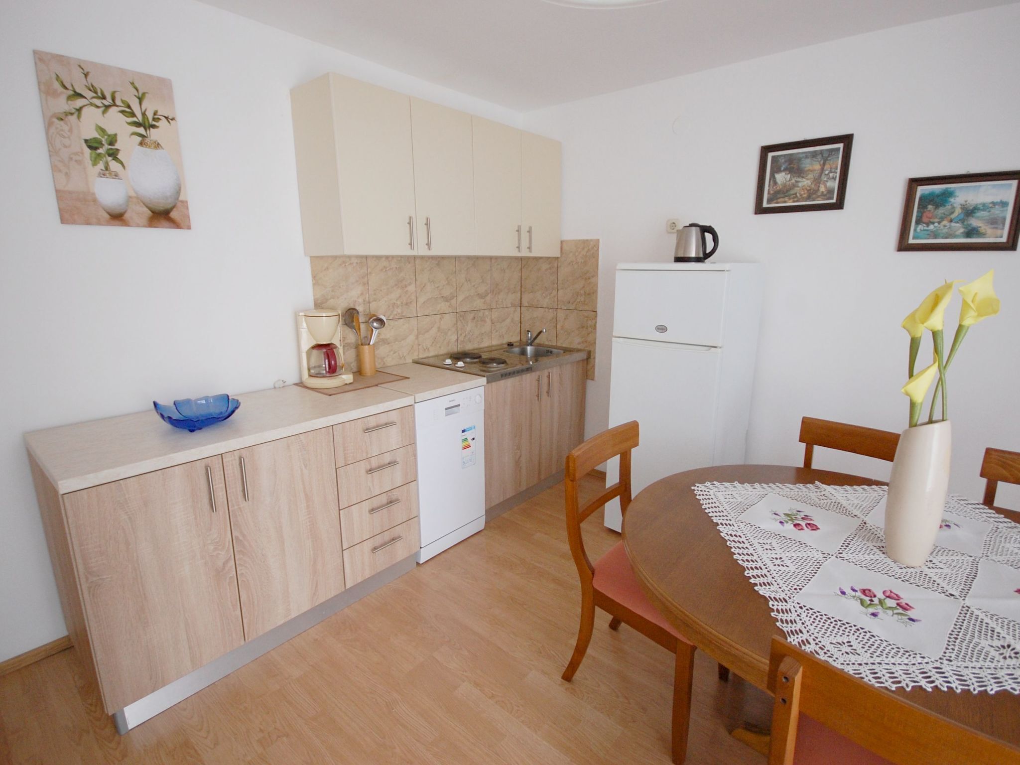 A compact kitchen with a dishwasher, a coffee maker, and a kettle on the counter. There is a dining table with chairs and a vase with flowers.