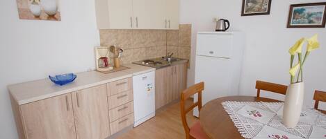 A compact kitchen with a dishwasher, a coffee maker, and a kettle on the counter. There is a dining table with chairs and a vase with flowers.