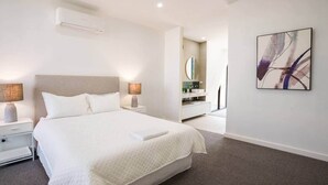 Ett modernt hotellrum med en stor säng, sänglampor, en väggmonterad luftkonditioneringsanläggning, ett badrum med en spegel och ett abstrakt konstverk på väggen.