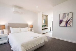 Ein modernes Hotelzimmer mit einem großen Bett, Nachttischlampen, einer wandmontierten Klimaanlage, einem Badezimmer mit Spiegel und einem abstrakten Kunstwerk an der Wand.