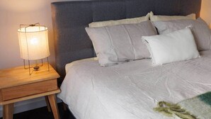 A bed with a grey headboard, a bedside table with a lamp, and a blanket on the bed. - Ocean-View Apartment in Heart of Portarlington (Portarlington)