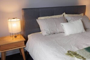 Ein Bett mit grauem Kopfteil, ein Nachttisch mit Lampe und eine Decke auf dem Bett.