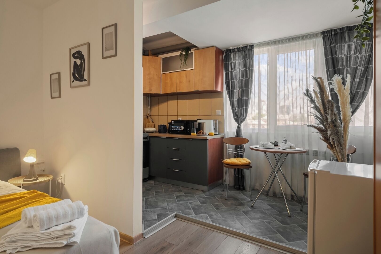 A compact hotel room with a bed, a small bedside table with a lamp, a kitchenette with a microwave, and a dining area with a table and chairs.