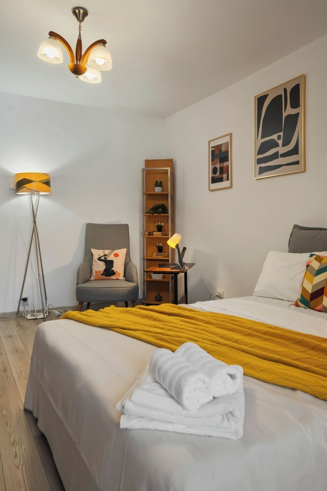 A hotel room with a bed, a desk, a chair, a floor lamp, a bookshelf, and artwork on the walls.