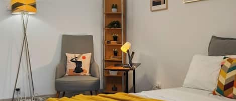 A hotel room with a bed, a desk, a chair, a floor lamp, a bookshelf, and artwork on the walls.