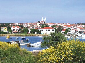 Une ville côtière où des bateaux sont amarrés le long de la côte, avec des bâtiments aux toits rouges et une tour imposante qui se détache à l’arrière-plan.