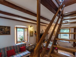 Ein rustikaler Innenraum mit freiliegenden Holzbalken, einer Treppe, einer Couch mit gemusterten Kissen sowie einem Essbereich mit Tisch und Stühlen.