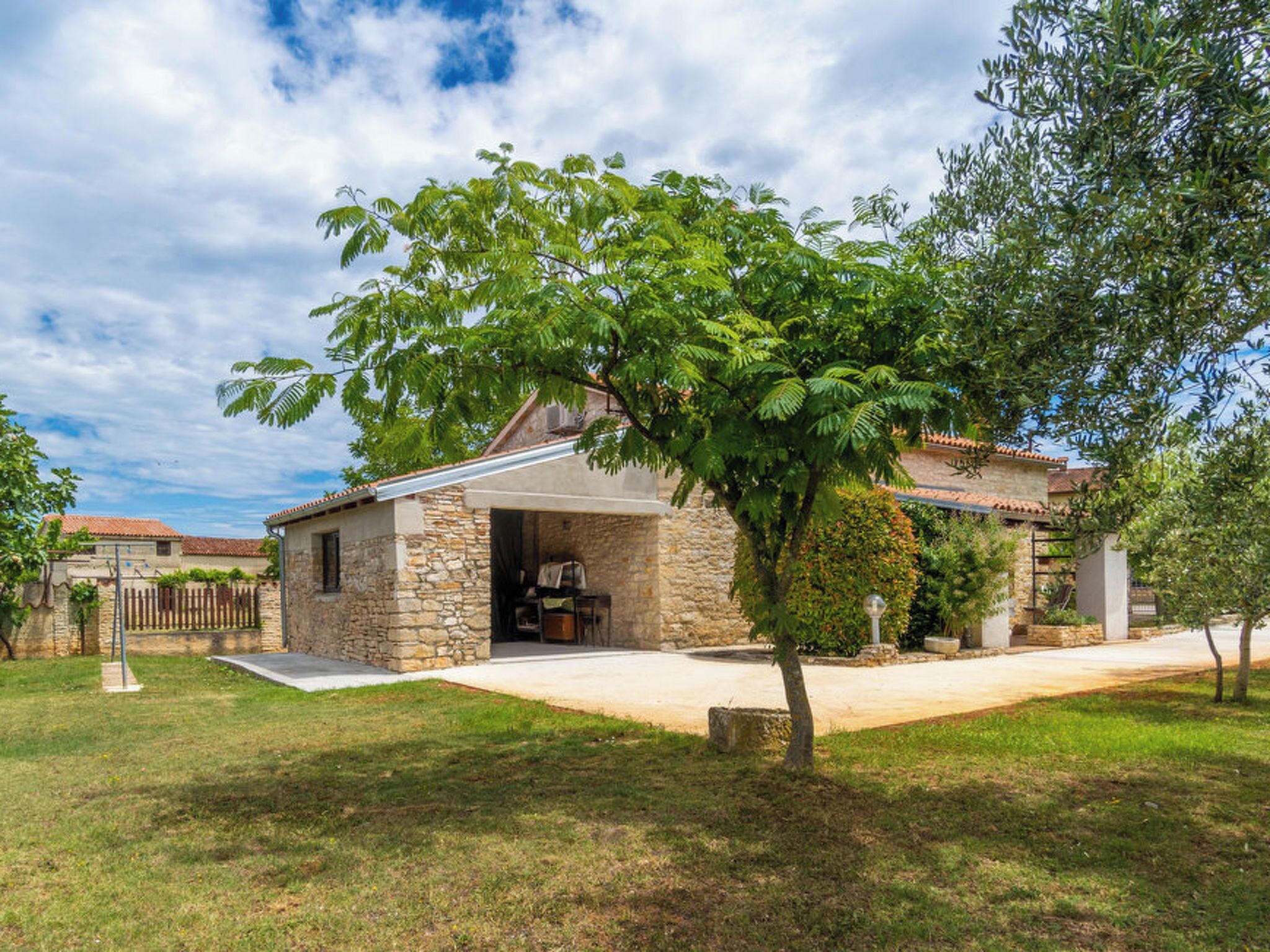 Une maison en pierre dotée d’un porche abrité, entourée d’une cour verdoyante et d’arbres.