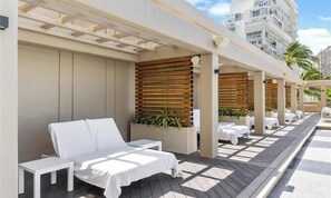 A sunlit outdoor seating area with white lounge chairs and tables, wooden slats, and potted plants. - Beautiful Studio at Ala Moana 1330 HONOLULU (Honolulu)