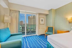 Une chambre d’hôtel avec un grand lit, une moquette à rayures bleues, un coin salon avec un fauteuil, une table et une vue sur de hauts immeubles, visible à travers une porte vitrée.