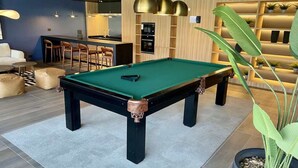 A pool table in a modern room with a bar area, wooden shelving with plants, and a seating area with a sofa and chairs. - Serviced Studio in Santiago in Greystar Somma (Santiago)