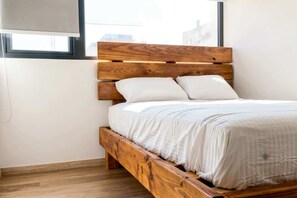 A bed with a wooden headboard and white bedding in a room with a window. - Furnished 2 Bedroom in Santiago in Greystar Somma (Santiago)