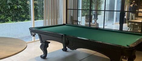 A billiards table in a room with large windows, a chandelier, and a view of greenery outside.