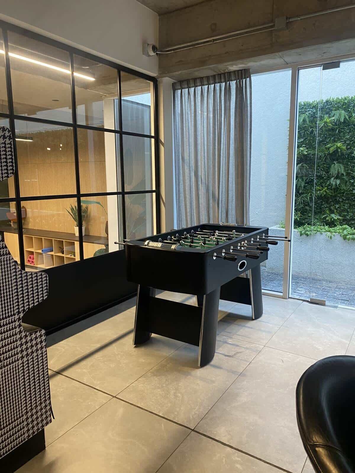 Een kamer met een tafelvoetbaltafel, een bank en een groot raam met gordijnen.