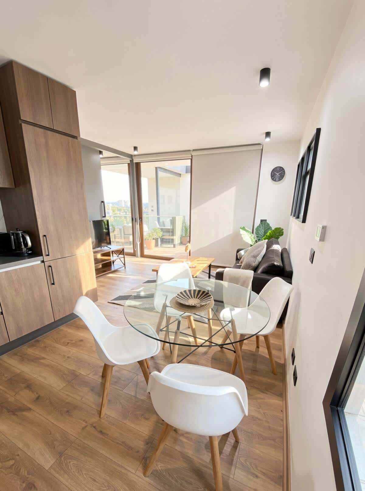 Une cuisine moderne avec des meubles en bois, un coin repas avec une table en verre et des chaises blanches, et un espace de vie avec un canapé et une plante.