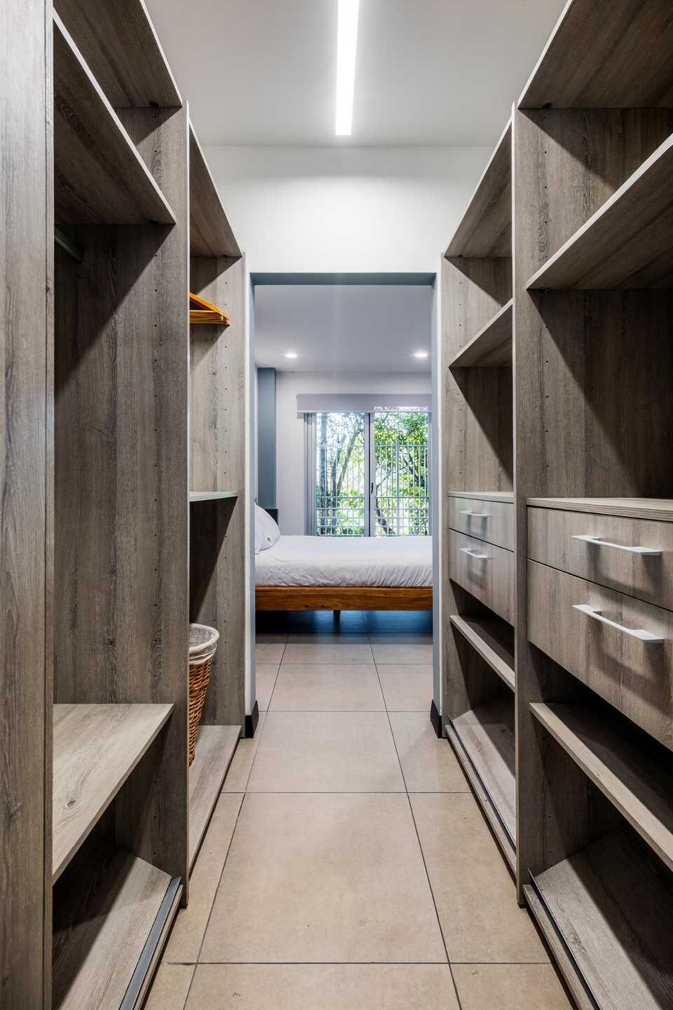 A narrow hallway with wooden shelving units on either side, leading to a bedroom with a bed and a window.