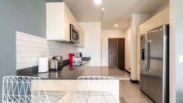 A modern kitchen with a stainless steel refrigerator, a microwave, and a toaster on the countertop. There are two white wire chairs and a small table in the foreground.