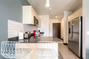 A modern kitchen with a stainless steel refrigerator, a microwave, and a toaster on the countertop. There are two white wire chairs and a small table in the foreground. - Cozy Studio in San Jose Urban Flats Building (Pozos)
